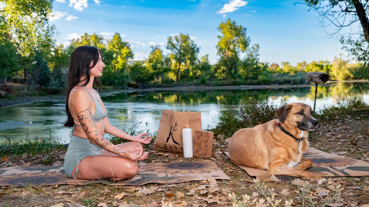 Woman meditating in lotus on a floral cork yoga mat beside a creek, her dog resting on a second cork mat, with a Kaya cork tote, water bottle and floral cork block between them